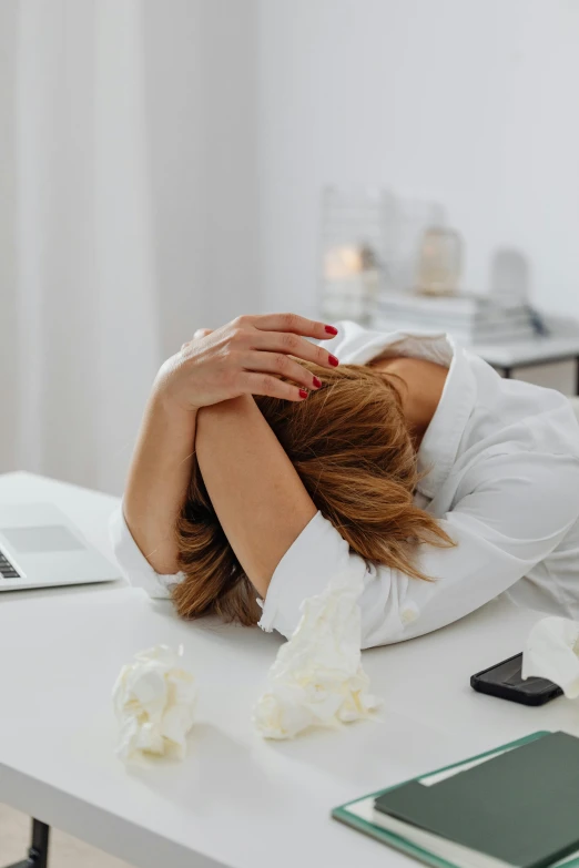 a woman sitting at a desk with her head in her hands, pexels, wilted flowers, in a white room, sitting at a computer, tired and drunk