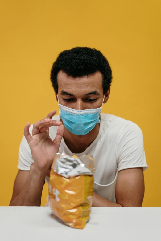 a man sitting at a table with a bag of food in front of him, pexels, hyperrealism, surgical mask covering mouth, yellow, ashteroth, pills