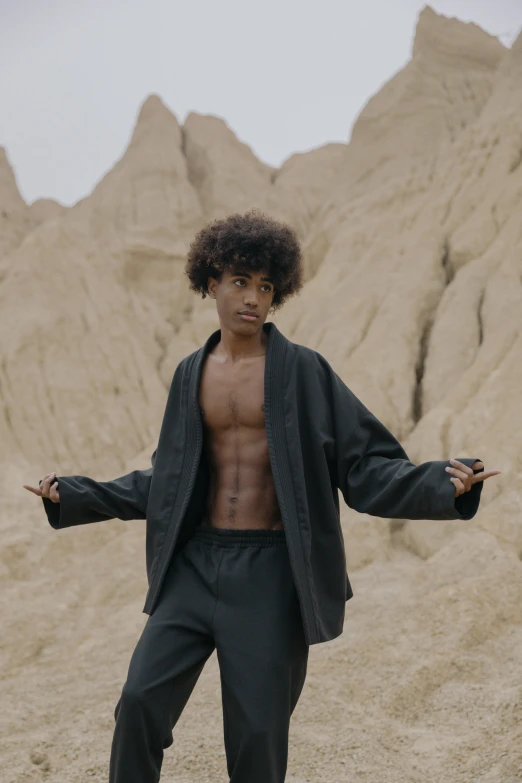 a man standing on a skateboard in the desert, an album cover, by Attila Meszlenyi, unsplash, afrofuturism, pretty samurai with afro, jacket over bare torso, black teenage boy, grey robes