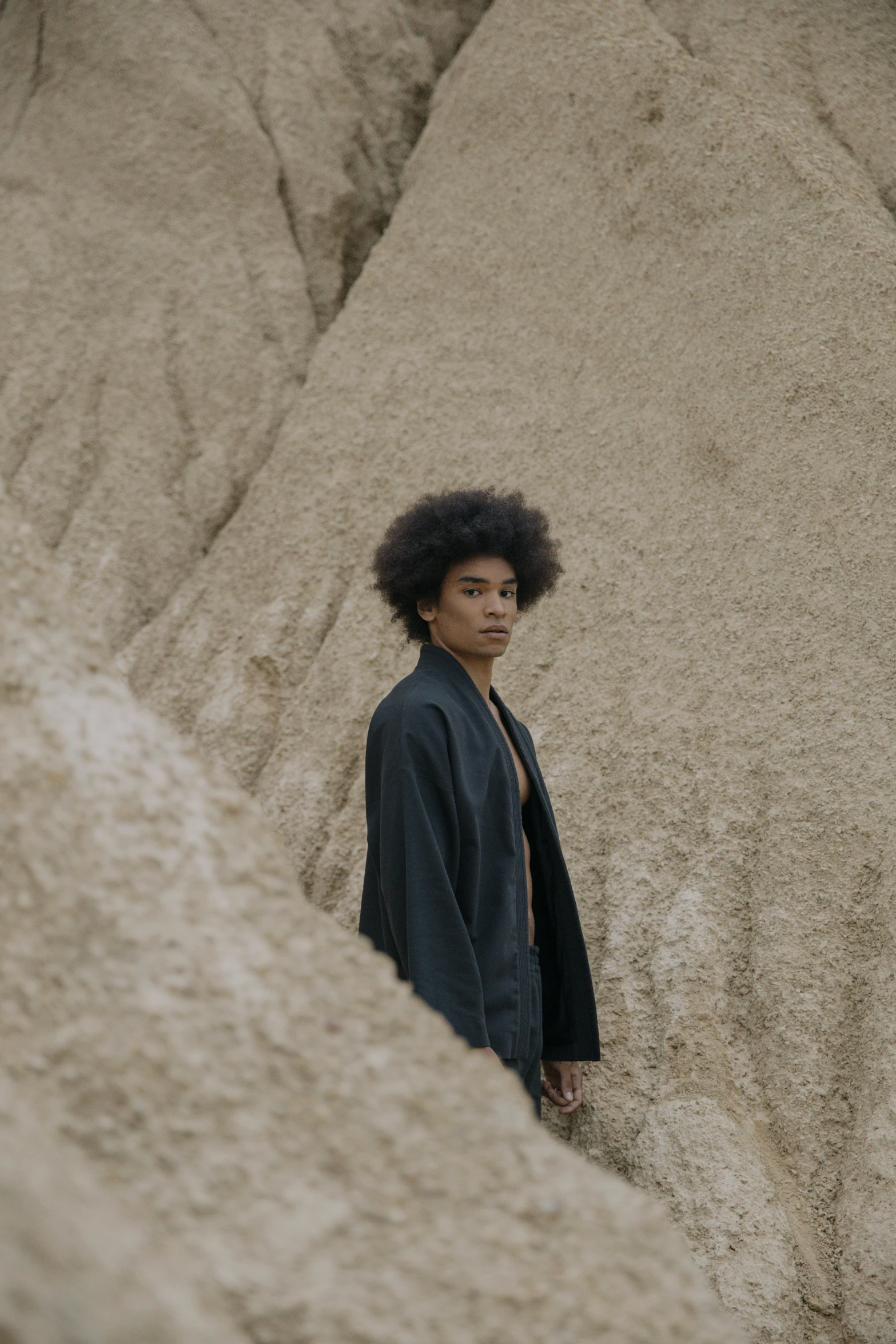 a man standing in front of a pile of sand, trending on unsplash, afrofuturism, wearing black silk robe, natural hair, portrait of a japanese teen, mountainside