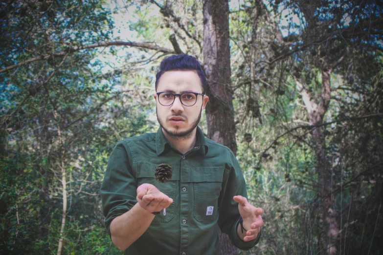 a man in a forest with a pine cone in his hand, an album cover, by Alejandro Obregón, pexels contest winner, hurufiyya, jewish young man with glasses, shrugging, instagram picture, low quality photo