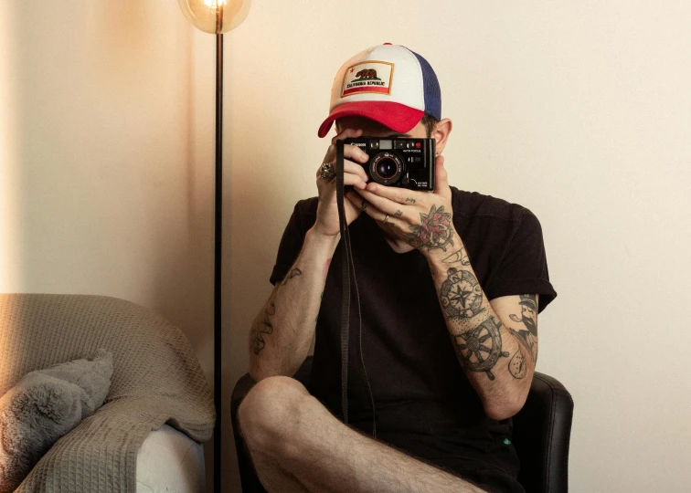 a man sitting in a chair taking a picture with a camera, a tattoo, red cap, h3h3, multicoloured, photography]