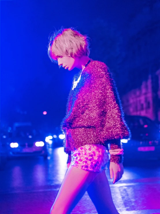 a woman walking across a street at night, an album cover, unsplash, happening, short platinum hair tomboy, patterned clothing, sequins, pink and blue lighting