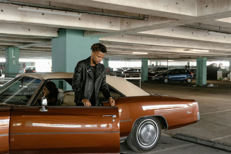 a man standing next to a car in a parking garage, an album cover, pexels contest winner, wearing a brown leather coat, standing in front of lowrider, in style of tyler mitchell, ( ( theatrical ) )