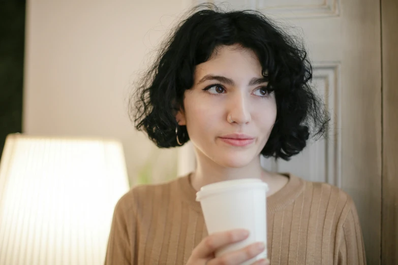 a woman holding a cup of coffee in her hand, a character portrait, trending on pexels, curly middle part haircut, pale-skinned persian girl, low quality photo, human staring blankly ahead