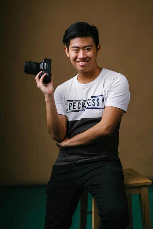 a man sitting on a stool holding a camera, a picture, inspired by Ryan Yee, headshot and bodyshot, slightly smiling, half asian, photography]