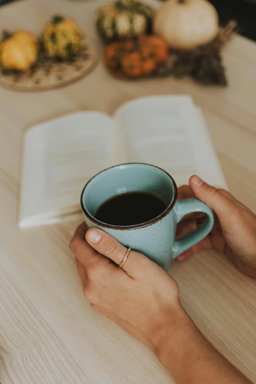 a person sitting at a table with a book and a cup of coffee, comforting and familiar, black and teal paper, connection rituals, early morning