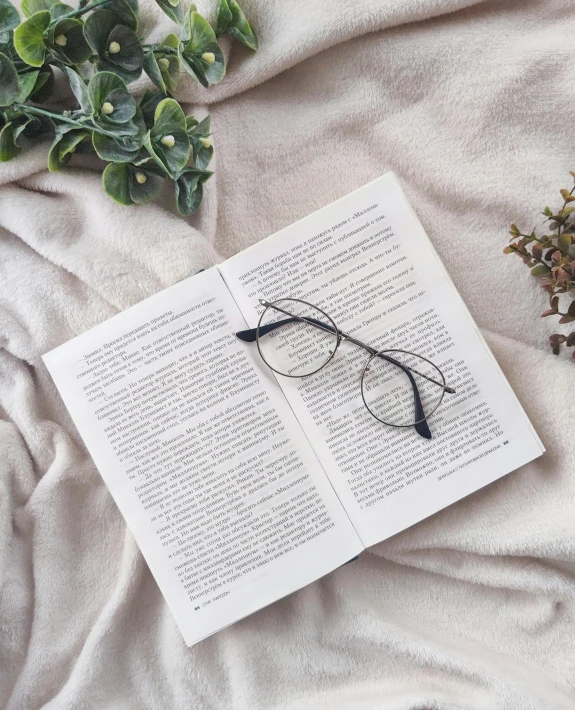 an open book sitting on top of a bed next to a plant, by Carey Morris, unsplash contest winner, square glasses, wearing a grey robe, high angle close up shot, textile