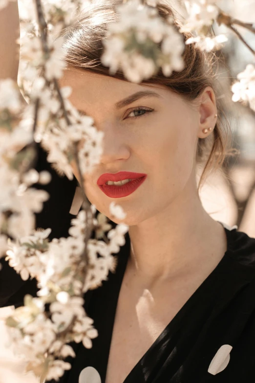 a woman standing in front of a tree with white flowers, a picture, by Mathias Kollros, trending on pexels, lipstick, chaumet, red, 15081959 21121991 01012000 4k