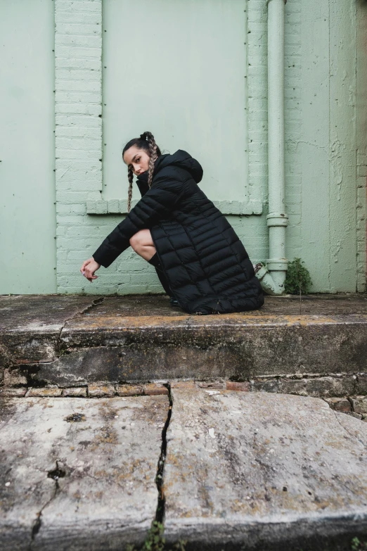 a woman sitting on the steps of a building, inspired by Elsa Bleda, unsplash, model wears a puffer jacket, battle pose, long black jacket, squatting