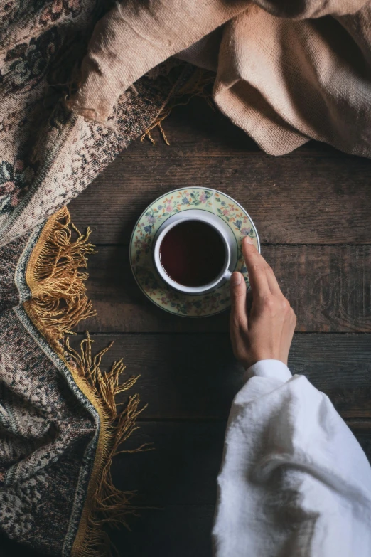 a person holding a cup of coffee on a table, a still life, trending on unsplash, hurufiyya, ornate flowing robe, assam tea garden setting, cozy calm! fabrics textiles, promo image