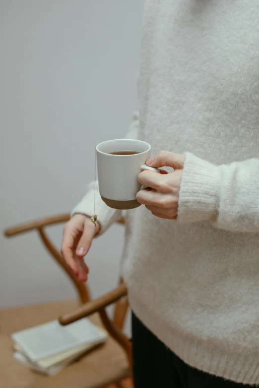 a woman in a white sweater holding a cup of coffee, by James Morris, trending on unsplash, minimalism, seated on wooden chair, tea cup, drinking cough syrup, on a white table