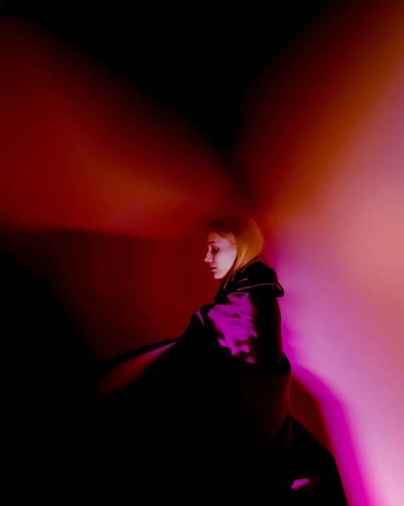 a woman standing in front of a pink light, an album cover, by Julia Pishtar, unsplash, darkness aura red light, wearing black clothes and cape, powerful woman sitting in space, looking off to the side