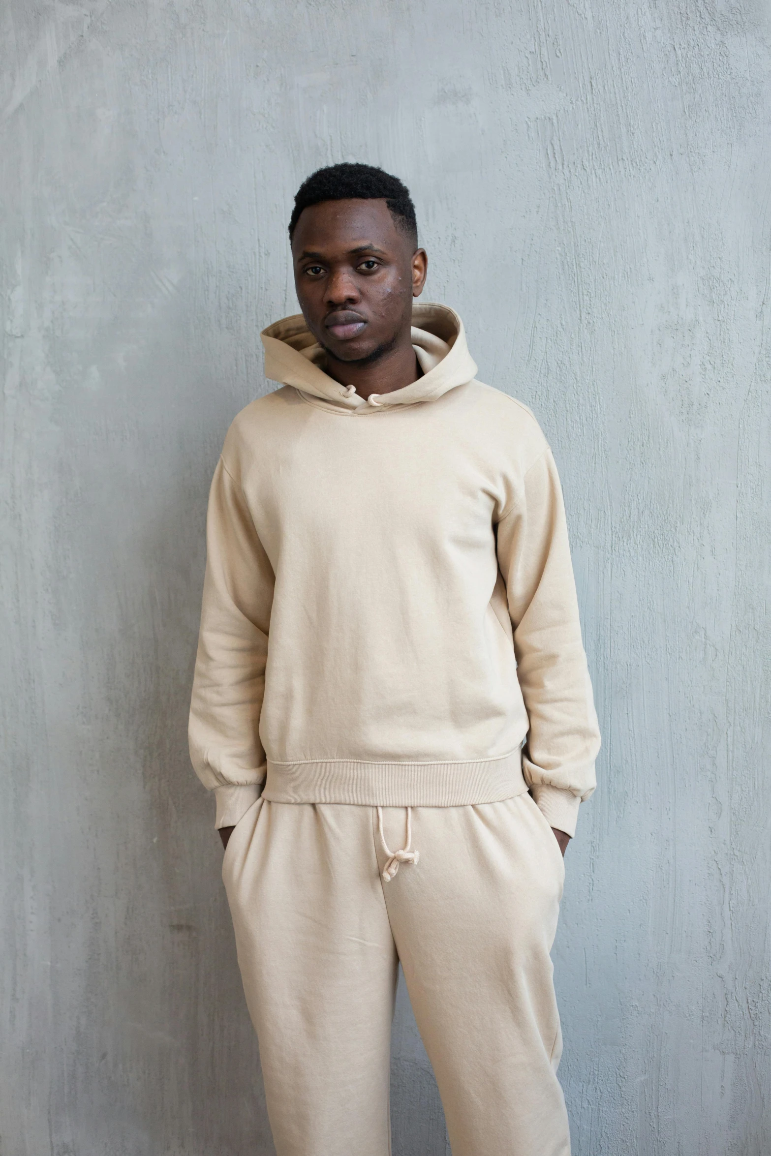 a man standing in front of a gray wall, an album cover, inspired by Barthélemy Menn, beige hoodie, wearing track and field suit, round-cropped, thumbnail