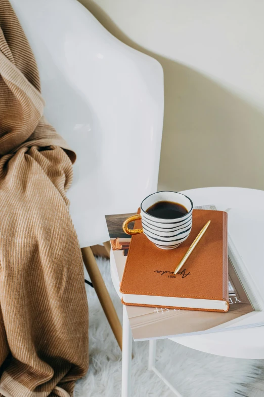 a coffee cup sitting on top of a white table next to a stack of books, brown clothes, orange robe, luxury journal cover, muted brown yellow and blacks