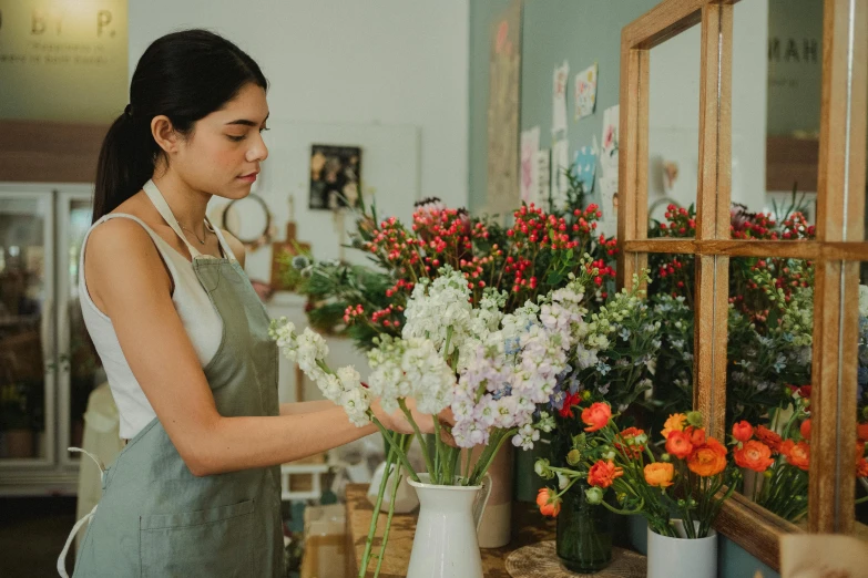 a woman is arranging flowers in a vase, trending on unsplash, arts and crafts movement, shop front, artist wearing overalls, thumbnail, attractive photo