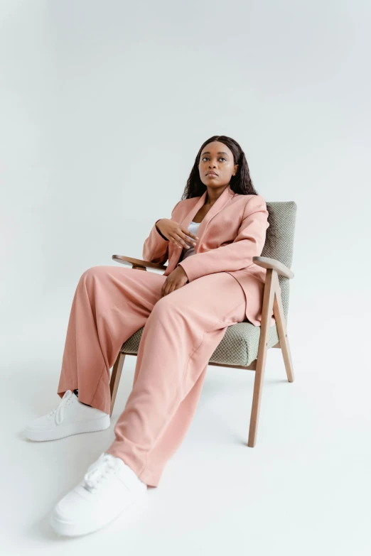 a woman in a pink suit sitting in a chair, by Maud Naftel, trending on unsplash, tessa thompson inspired, large pants, wearing wool suit, set against a white background