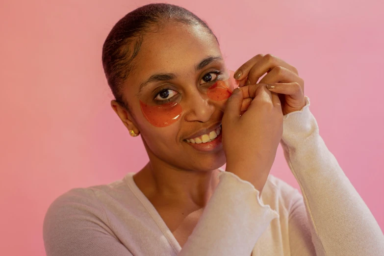 a woman putting makeup on her face in front of a pink background, pexels contest winner, process art, eyepatches, light-brown skin, wearing translucent sheet, left eye red stripe