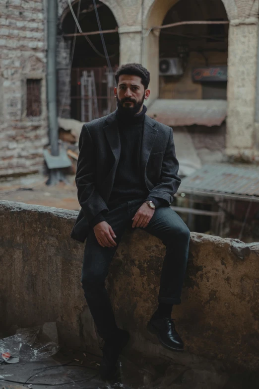 a man sitting on top of a stone wall, an album cover, pexels contest winner, a portrait of rahul kohli, wearing causal black suits, standing in township street, cinematic outfit photo