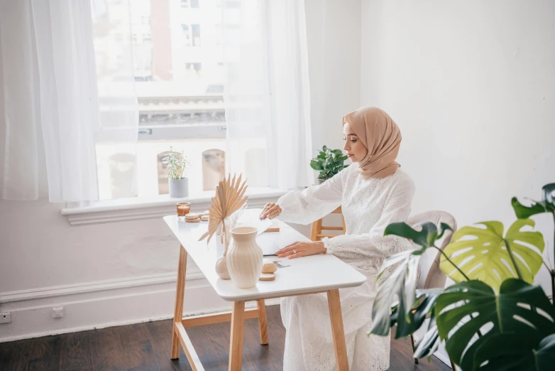 a woman sitting at a table in front of a window, inspired by Maryam Hashemi, pexels contest winner, hurufiyya, wearing white clothes, islamic interior design, tending on art station, sitting on a mocha-colored table