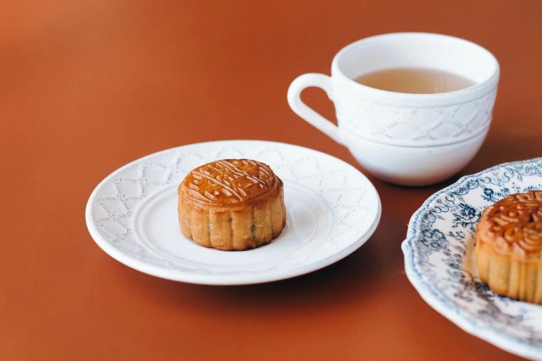 two pastries and a cup of tea on a table, inspired by Richmond Barthé, unsplash, hurufiyya, chinese empress, flan, on a pale background, hong kong