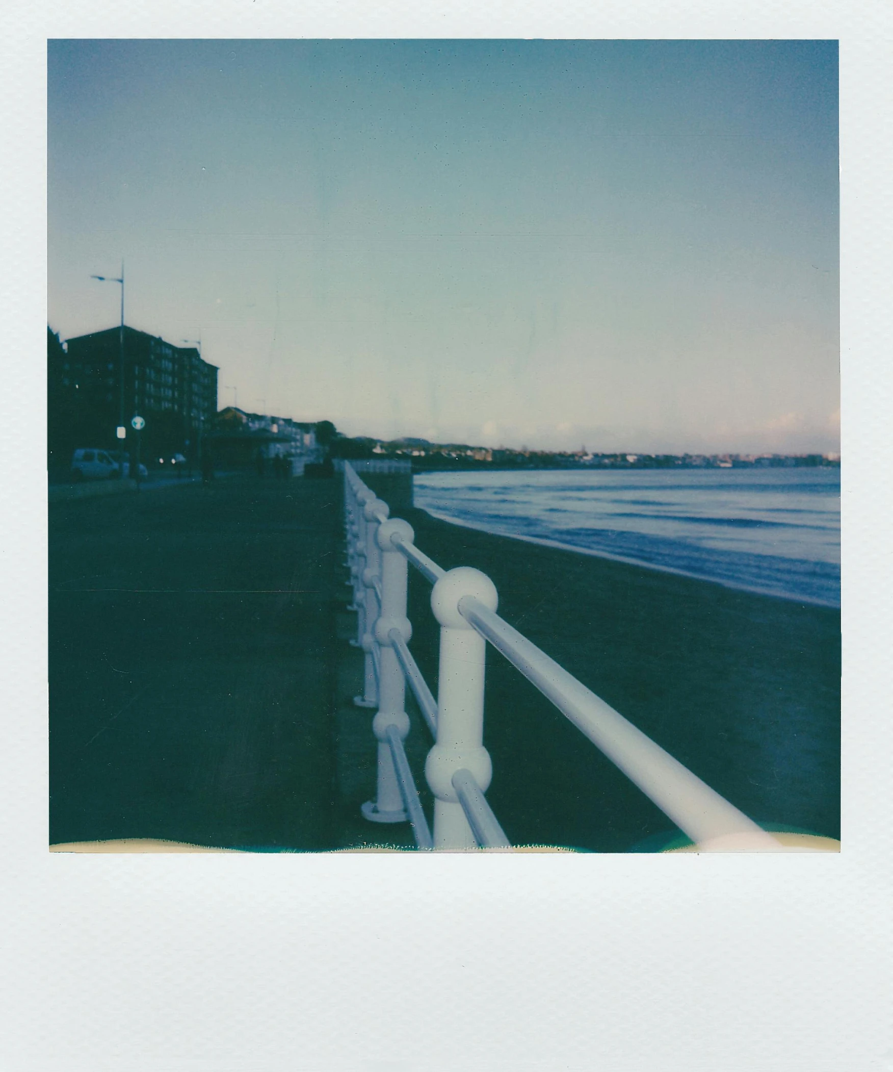 a white railing next to a body of water, a polaroid photo, the sea seen behind the city, polaroid colour faded, (night), beachfront