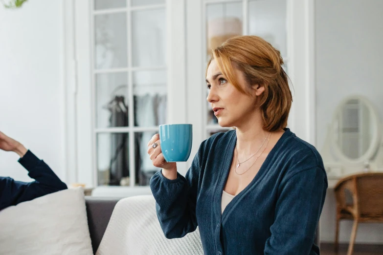 a woman sitting on a couch holding a cup of coffee, by Julian Hatton, pexels, hurufiyya, blue robe, worried, alison hannigan, seen from the side