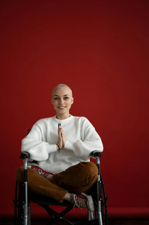 a woman sitting in a wheelchair in front of a red wall, shaved head, anjali mudra, healthcare, asher duran