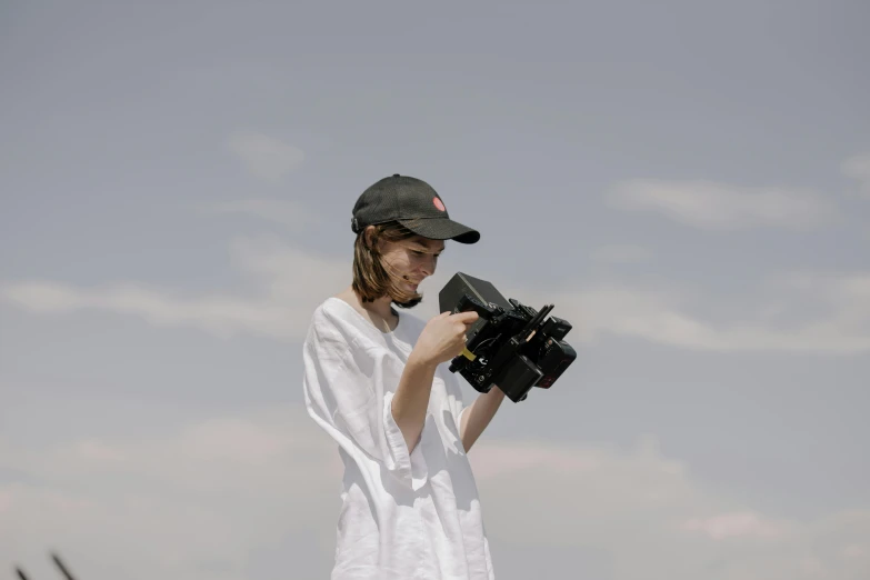 a woman standing in a field holding a camera, video art, uniform off - white sky, wearing sunglasses and a cap, medium format, portait image