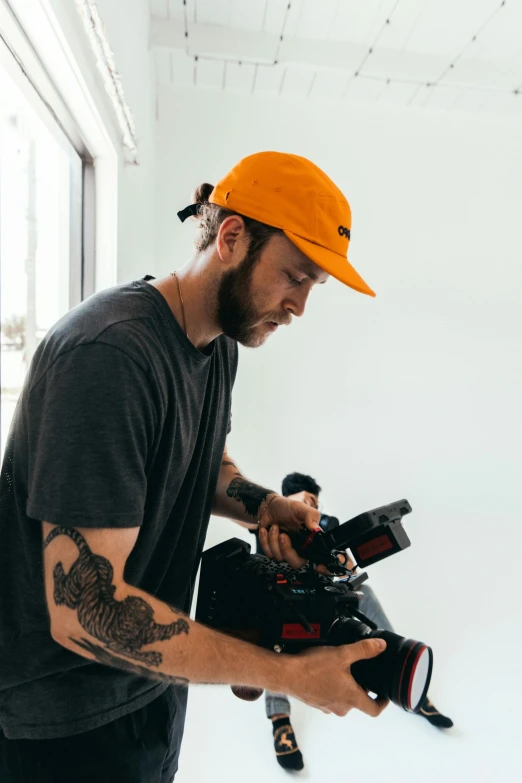 a man holding a camera in a white room, wearing a red backwards cap, dylan kowalsk, screenwriter, tattooed