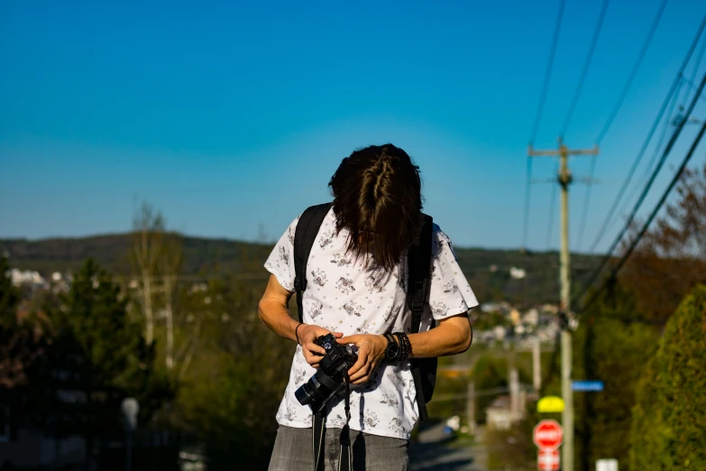a man standing on the side of a road holding a camera, by Julia Pishtar, happening, on a bright day, student, shot from the back, mixed art