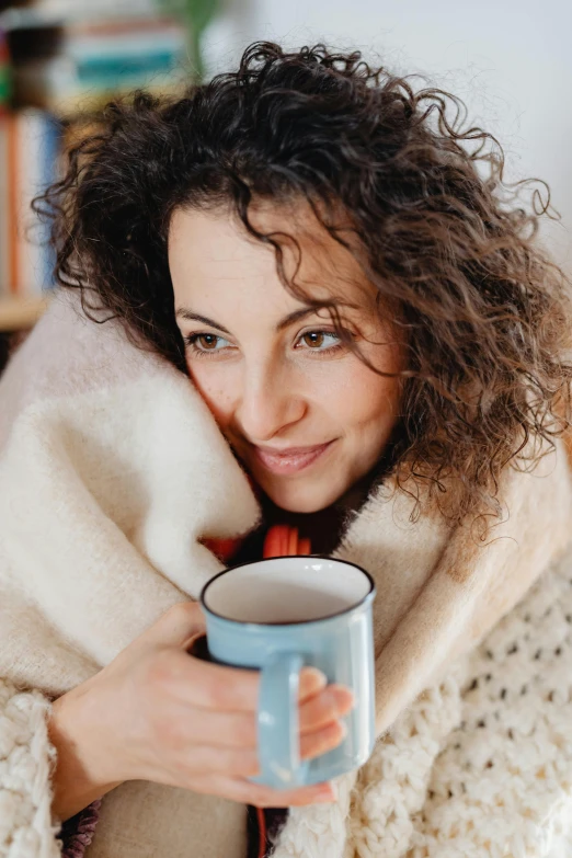 a woman wrapped in a blanket holding a cup of coffee, a portrait, trending on pexels, brown curly hair, blue, square, contented