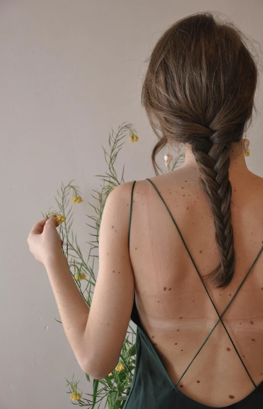 a woman standing in front of a vase of flowers, a tattoo, inspired by Elsa Bleda, trending on pexels, braided brown hair, white freckles, back - lit, wearing a camisole