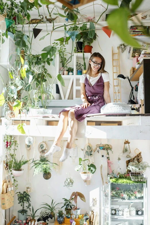 a woman sitting on top of a desk in a room filled with plants, inspired by Yuko Tatsushima, trending on pexels, plants in glasses, in a white boho style studio, artist wearing overalls, high angle