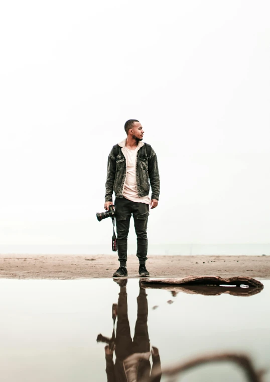 a man standing on a beach next to a body of water, by Robbie Trevino, an aviator jacket and jorts, full body camera shot, transparent background, reflective ground