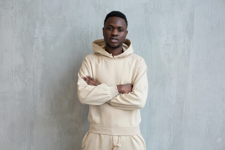 a man standing with his arms crossed in front of a wall, inspired by Paul Georges, trending on pexels, beige hoodie, maria borges, tracksuit, press shot