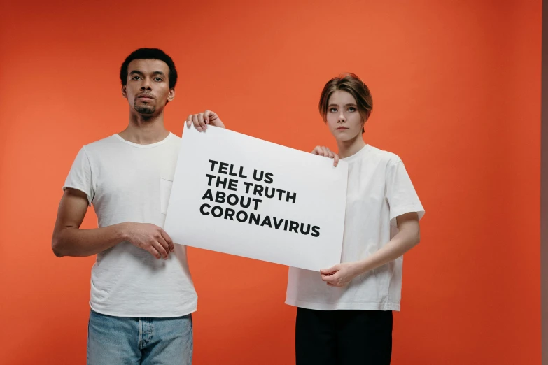 two people holding a sign that says tell us the truth about coronavirus, pexels contest winner, orange and white, caparisons, biological photo, looking partly to the left