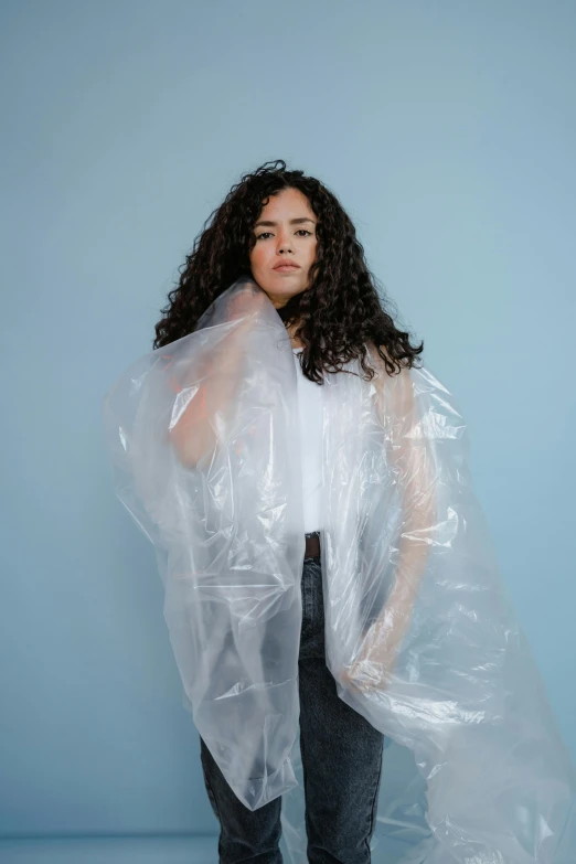 a woman in a plastic raincoat standing against a blue wall, by Olivia Peguero, curly long hair, wearing a plastic garbage bag, on a gray background, wearing a king's cape