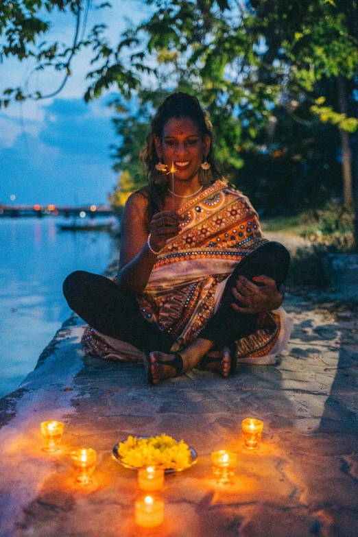 a woman sitting on a wall with candles in front of her, with lotus flowers, dark skinned, sitting in front of a lake, celebrating