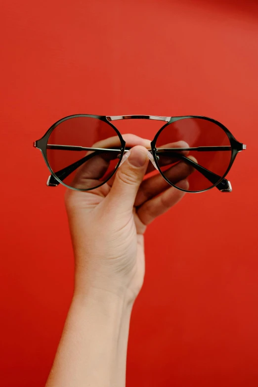 a hand holding a pair of glasses against a red wall, by Sven Erixson, pexels contest winner, photorealism, black sunglasses, square rimmed glasses, black on red, seventies