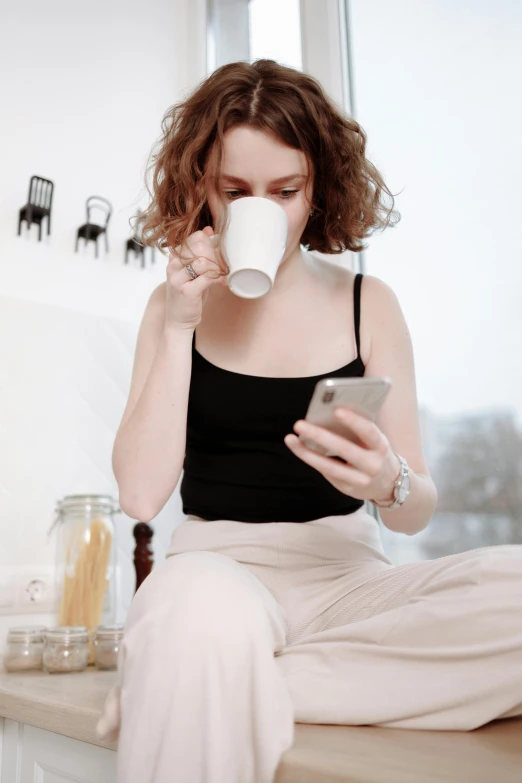 a woman sitting on a counter using a cell phone, trending on pexels, is ((drinking a cup of tea)), pale skin curly blond hair, long coffee brown hair, large)}]