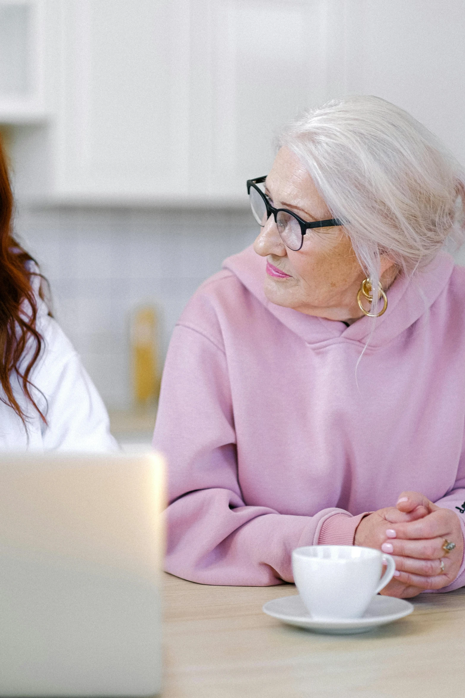 two women sitting at a table in front of a laptop, a digital rendering, trending on pexels, white-haired, pondering, nursing, background image