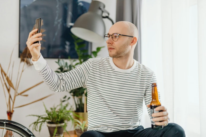 a man sitting on a bed holding a beer and a cell phone, pexels contest winner, no hair completely bald, square rimmed glasses, avatar image, mirror selfie