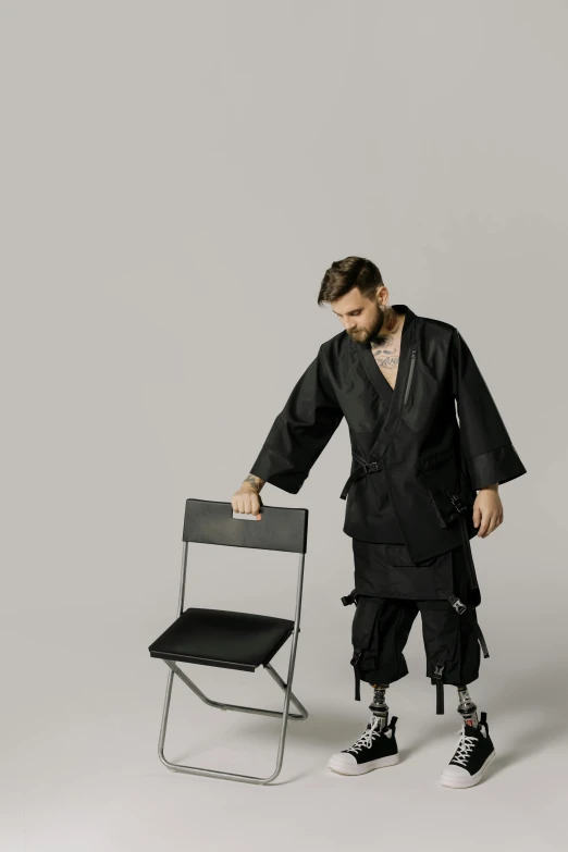 a man standing next to a folding chair, by Nina Hamnett, purism, karate outfit, black reflect robe, post malone, fighter pose