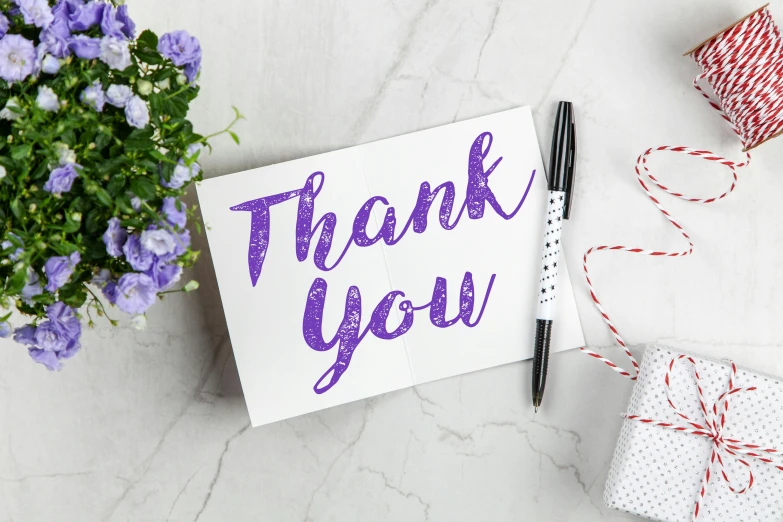 a thank card sitting on top of a table next to a bunch of flowers, by Elaine Hamilton, trending on pexels, graffiti, purple sparkles, pen on white paper, background image, angled view