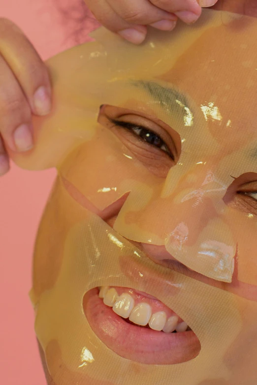 a woman is putting a mask on her face, by Ellen Gallagher, reddit, plasticien, candy treatments, gold skin, smiling mask, dynamic closeup