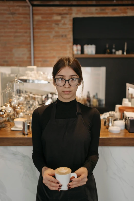 a woman standing in front of a counter holding a cup of coffee, round black glasses, asher duran, looking serious, ash thorp khyzyl saleem