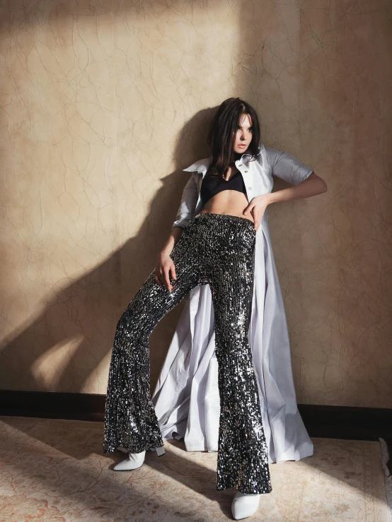 a woman standing in a room next to a wall, an album cover, by Nina Hamnett, trending on pexels, baroque, sequin top, wearing long silver robes, kendall jenner, breeches