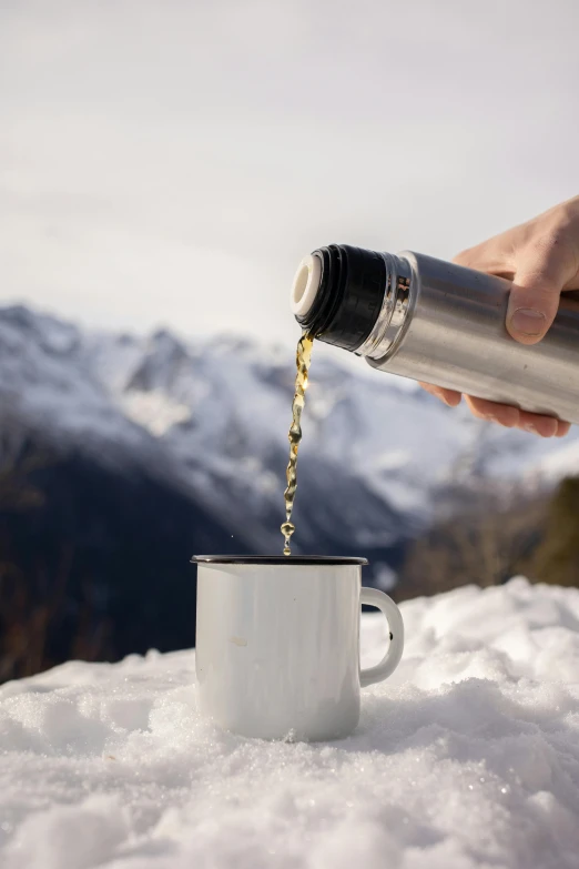 a person pouring wine into a cup in the snow, top lid, hot coffee, with mountains as background, stainless steel