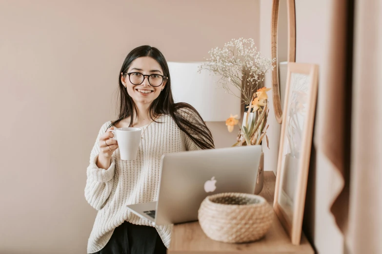 a woman holding a cup of coffee in front of a laptop, trending on pexels, in square-rimmed glasses, standing in corner of room, avatar image, smiling sweetly